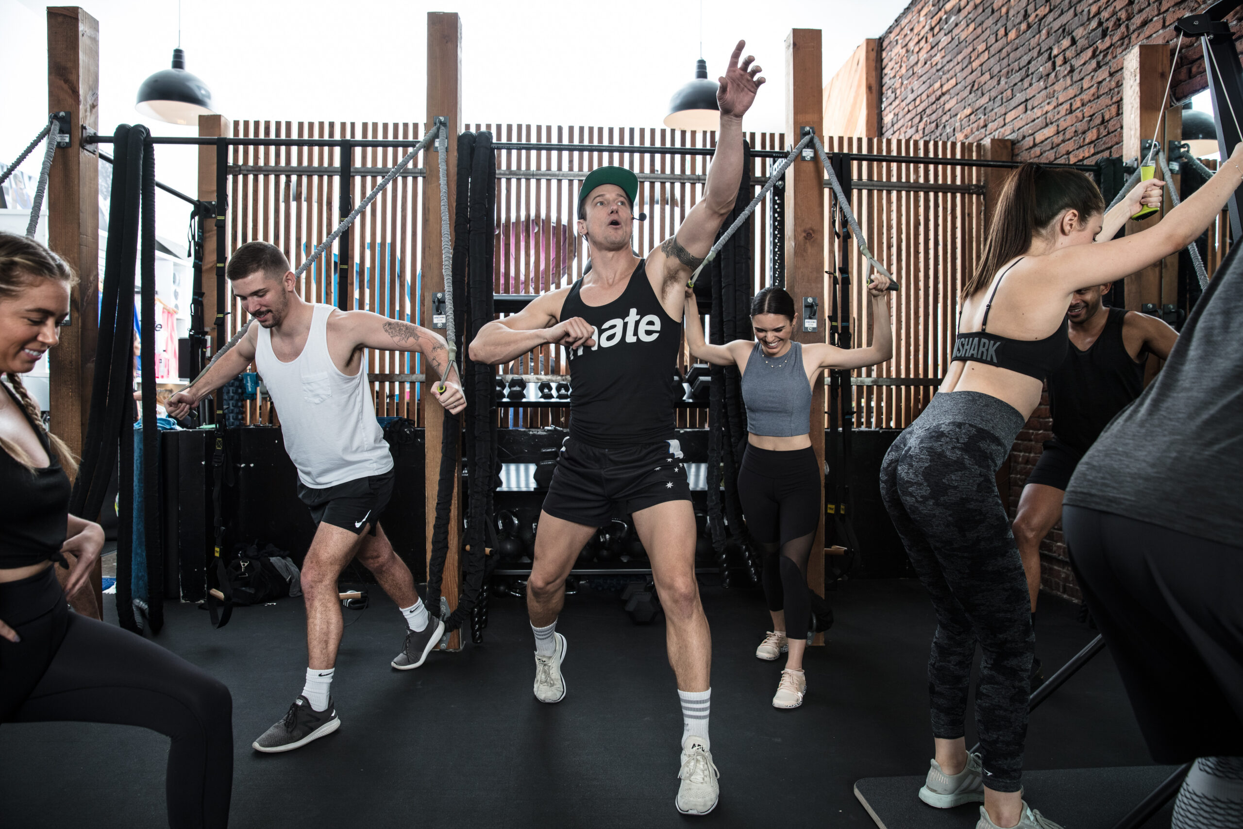 A high-energy Training Mate coach leading a group fitness circuit, demonstrating proper form while participants engage in resistance cable exercises in a modern, open-air studio